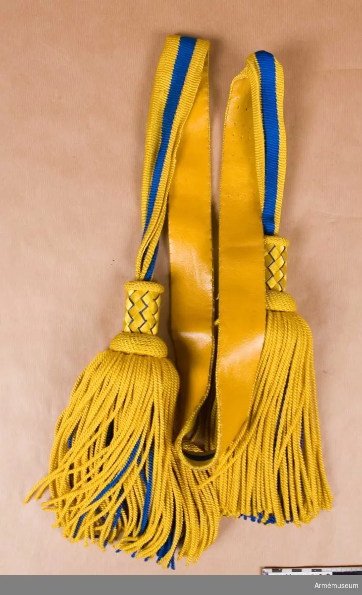 Skärp m/1819-1829 för kompaniofficer, A 1. Av silke, språngat med två gula och två blå ränder. Tofsar i ändarna med korgflätad  Överdel i blått och gult. Tofsen är i tvinnat silke, gult på  utsidan och blått innanför i samma grovlek och längd. Skärpet är fodrat med gult skinn.