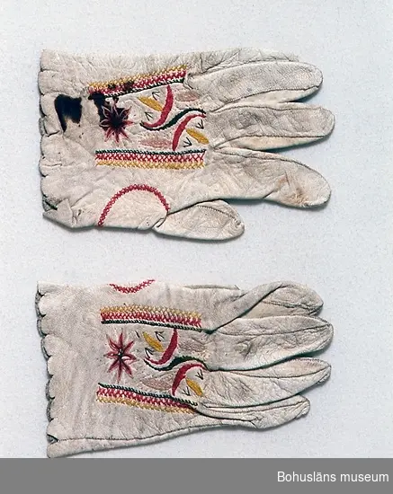 Handgjorda handskar av vitt skinn med broderi i bomullsgarn och silke i rött, ljust rosa, grönt, ljust gråbrunt och gultonat vitt.
Broderi i plattsöm, flätsöm och raka stygn finns på ovansidan. Sömmen runt tummen är markerad med en flätsöm.
Tillverkningstid och användningstid uppskattade. 
Vantarna är märkta UM4407 a och b.
Den blivande bruden fick så kallade brudhandskar som gåva av fästmannen. Hon skulle bära dem på bröllopet, men de användes även senare till högtid.
Slitna. Två stora bruna fläckar på ovansidan av ena handsken.
En del av broderiets tråd bortsliten. Smutsiga.

Litteratur:
Nylén, Anna-Maja, Folkdräkter ur Nordiska  museets samlingar, Nordiska museets handlingar 77, Lund 1971, sid. 67-70. 
Takhimlar och brudhandskar Folklig textiltradition i Västergötland, Widhja, Inger, Skrifter från Skaraborgs länsmuseum nr 12, Skara 1990, sid. 132-133.

Ur handskrivna katalogen 1957-1958:
Ett par brudhandskar
L. c:a 18 cm; vitt skinn m. broderi i ylle; ngt smutsiga, på b) två mörka? fläckar. Hela.
