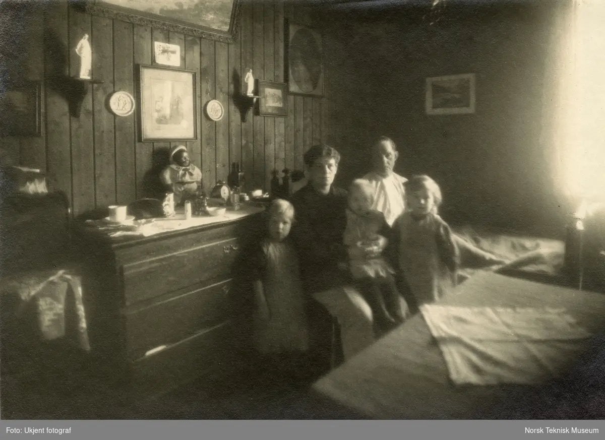 Mor og far med to barn fotografert i kombinert stue og soverom. Bilde brukt i Barselhjemustillingen 1916