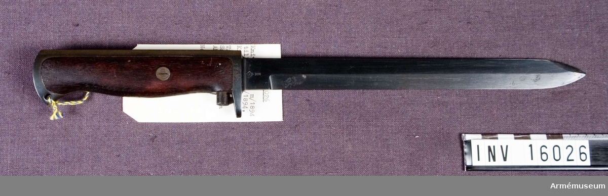 Samhörande nr är 16026-16027.Knivbajonett m/1894 till gevär m/1894, Krag Jörgensen, Norge.Klingans bredd vid fästet: 19 mm. Grepplattorna av trä fastsatta med skruv. Bajonetten  är smidd i ett stycke och har eneggad klinga. Låsknappen är placerad omedelbart ovanför parerstången och står i förbindelse med spärren i grepphuvudet via en stång som löper diagonalt genom greppet. Märkt: Kung Haakons namnchiffer 7. (1905-1957).  Tillv.nr: 72553.Samhörande: AM 16026 knivbajonett m/1894. AM 16027 balja av stålplåt till bajonett m/1894.