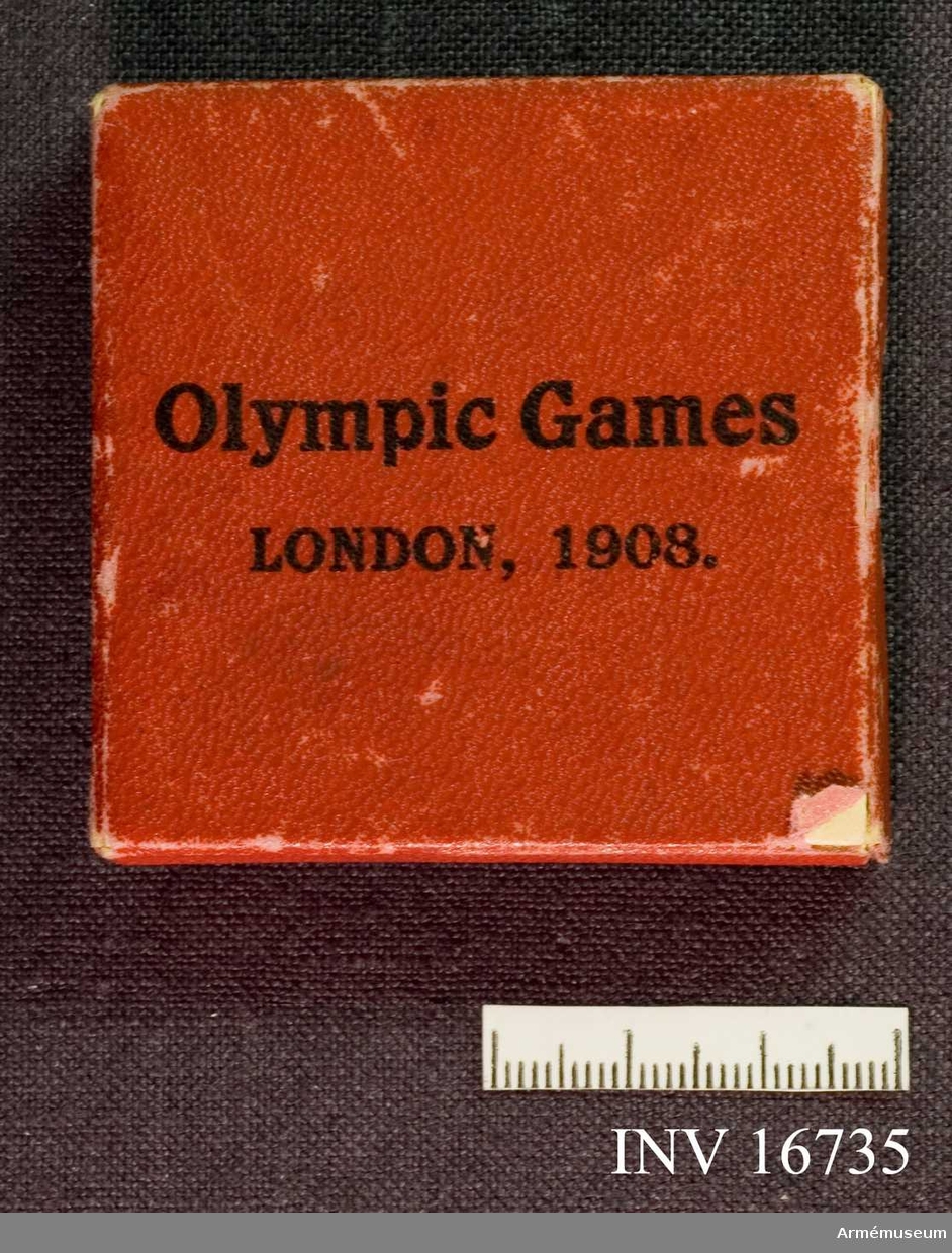 Minnesplakett från olympiska spelen i London 1908 av mattförsilvrad metall.

Åtsidan avbildar segergudinnan Nike, medan frånsidans motiv återger en scen med fyra hästar som drar en stridsvagn med körkarl och segerherre.

Plaketten är placerad i en fyrkantig röd ask.
