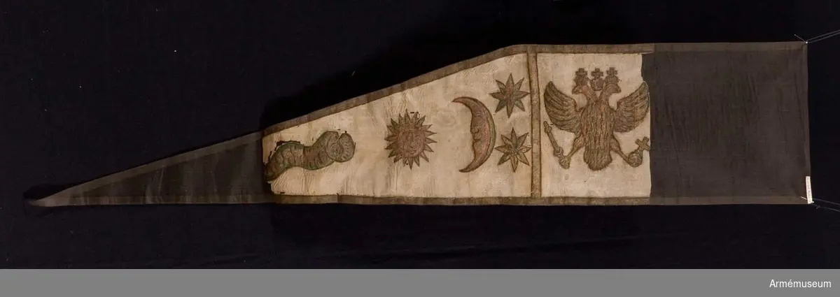 Duk av vit sidentaft. Motivet målat i guld- och silver samt rött och grönt bestående av en krönt örn, måne, sol och stjärnor, På duken en pappersetikett som anger att den tagits i slaget vid Saladen 1703.