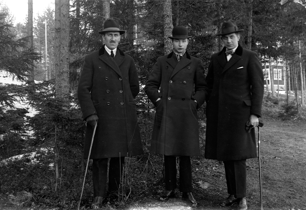 Tre män klädda i överrock, två har promenadkäpp i handen, Gustav Andersson (2). De står i skogen och i bakgrunden skymtar hus.