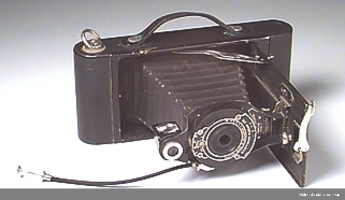 Svart hopfällbar kamera av märket HAWK-EYE model B. Avtryckarknappen sitter fästad på en 130 mm lång sladd. Handtag i mörkt läder märkt: No 2 A FOLDING CARTRIDGE HAWK EYE. Bredden i ihopfällt tillstånd är 87 mm. Bakstycket är märkt: USE FILM No 116 (använd film nr 116). 