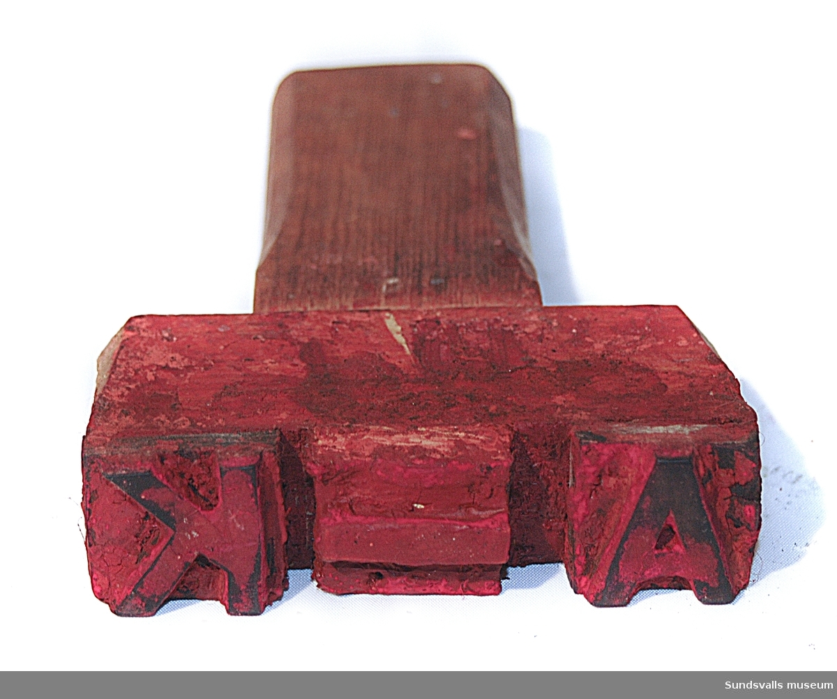 Stämpel i gummi med handtag i trä. Stämpeln visar bokstäverna A-K Röda färgrester finns kvar på stämpeln.