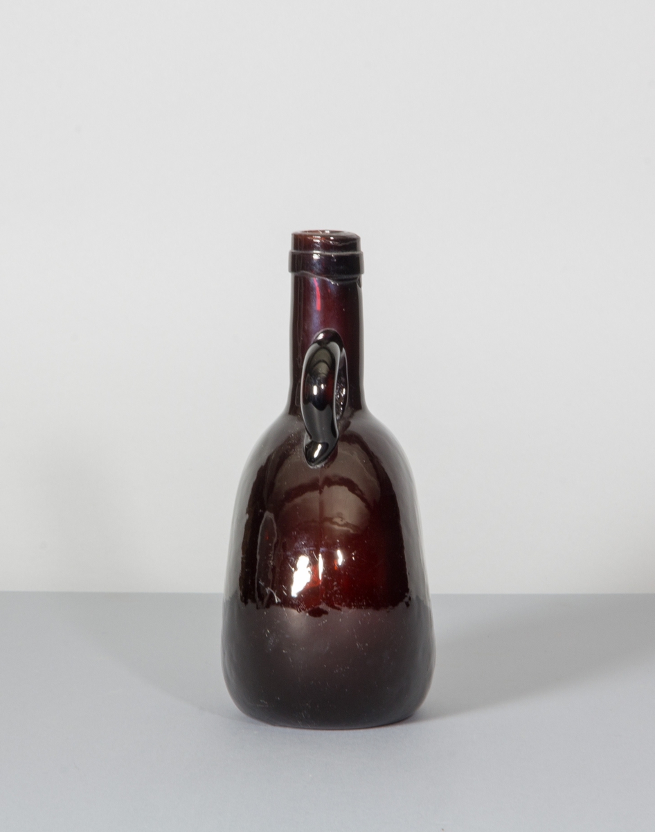 Flaska i rödbrunt glas. Tillplattad modell med rundade sidor. Hals samt påklippt hänkel.
Likheter med den tyska Bocksbeutel.