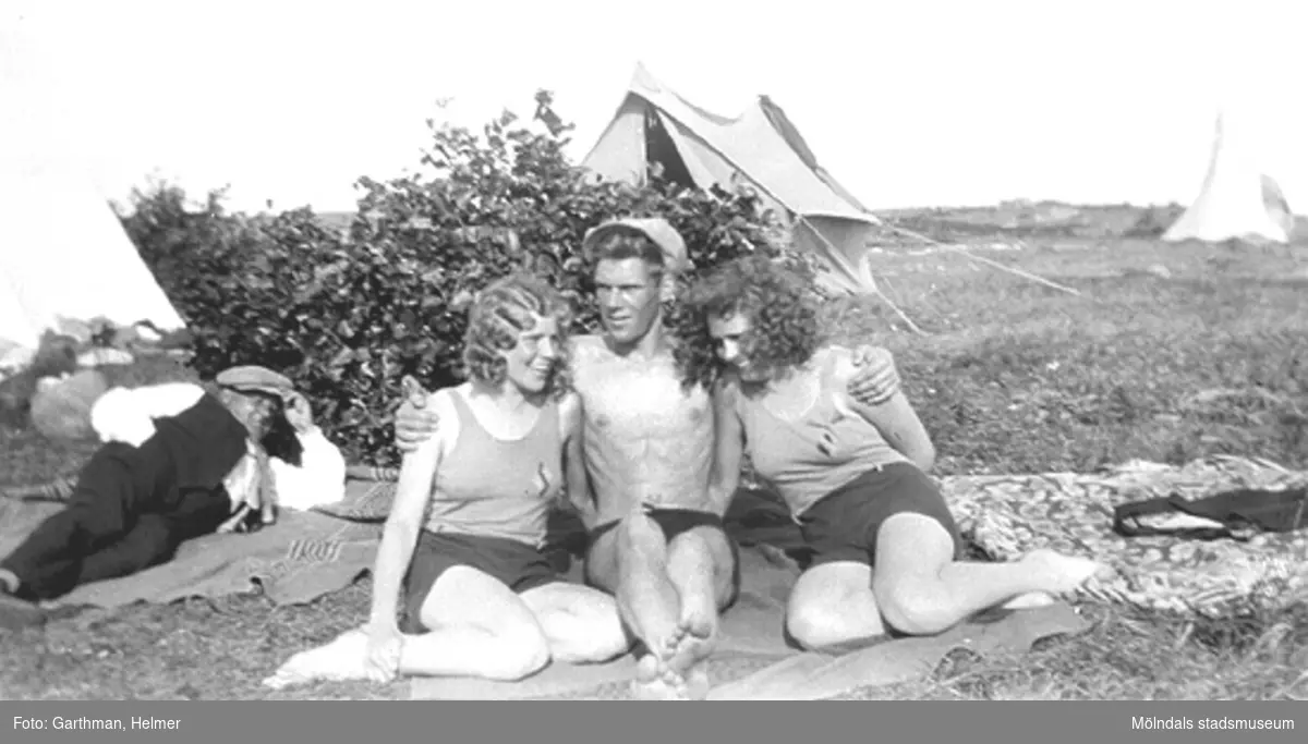 Helmer Garthmans kompisar, från vänster: Anna Nilsson, Wilgot och Gitt Ullberg
. De sitter på en filt framför en buske vid sjön Lygnern i Fjärås. Okänd man i bakgrunden. 1930-tal.
