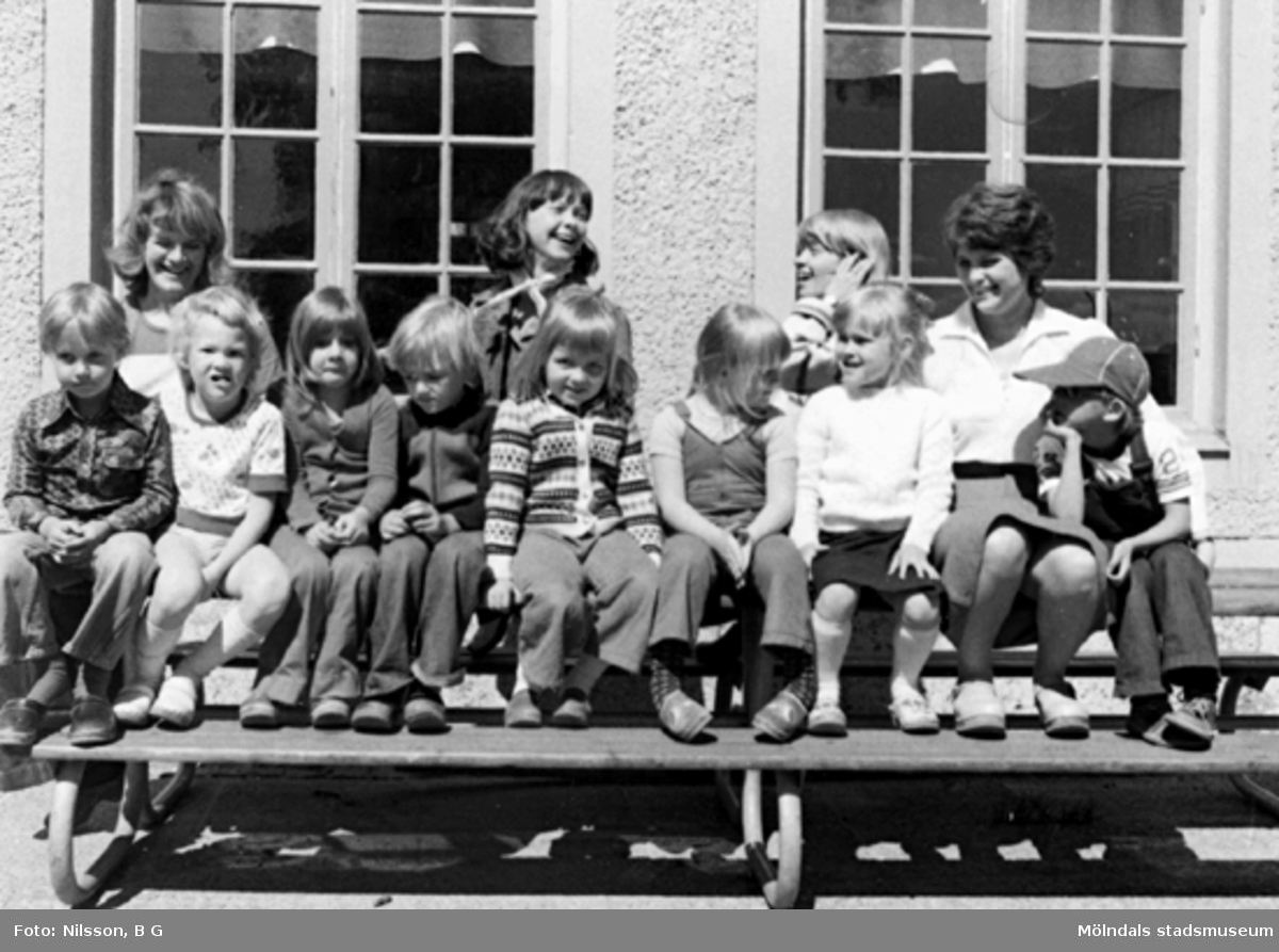 Barn och fröknar som sitter på en bänk utomhus. Holtermanska daghemmet, okänt årtal.