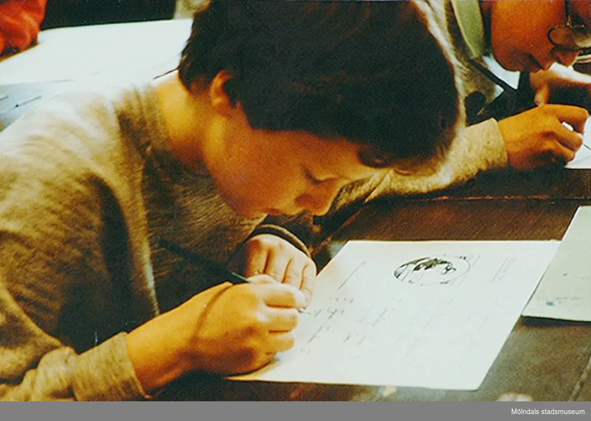 Ett barn som sitter vid en bänk och skriver finstil med bläckpenna. Okänt årtal.