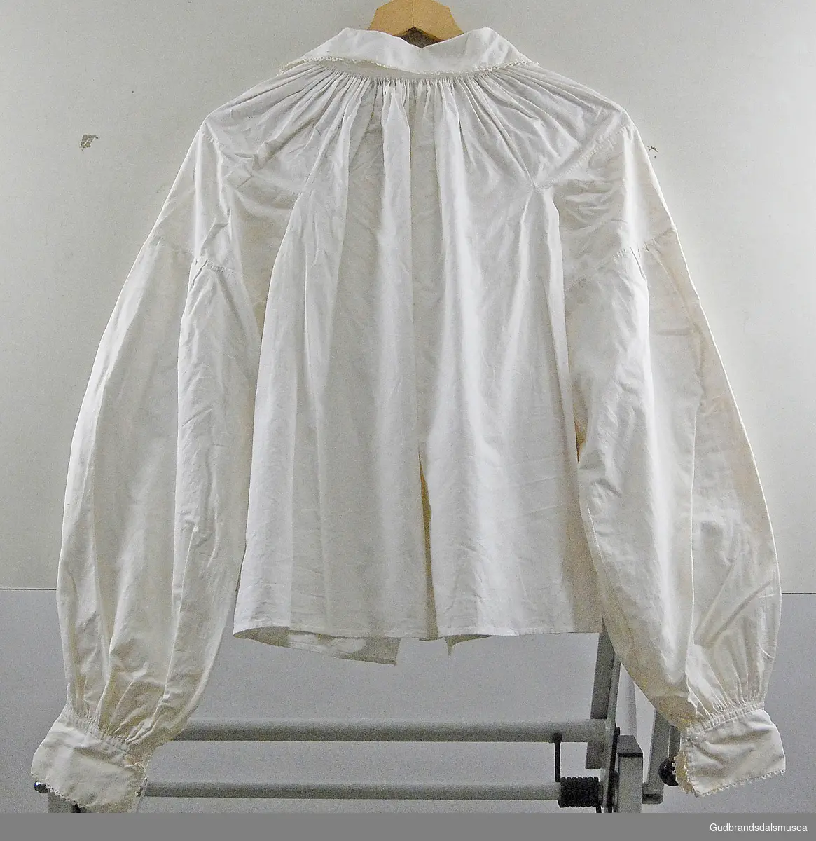 Hvit bunadsskjorte, dame, med nuperelle-kanter rundt nedre kanter på ermer og krage. Folder bak og foran (på/ved nakke), det samme ved ermekanter. Har "doble" plastknapper/"plastmansjetter". Åpning foran.