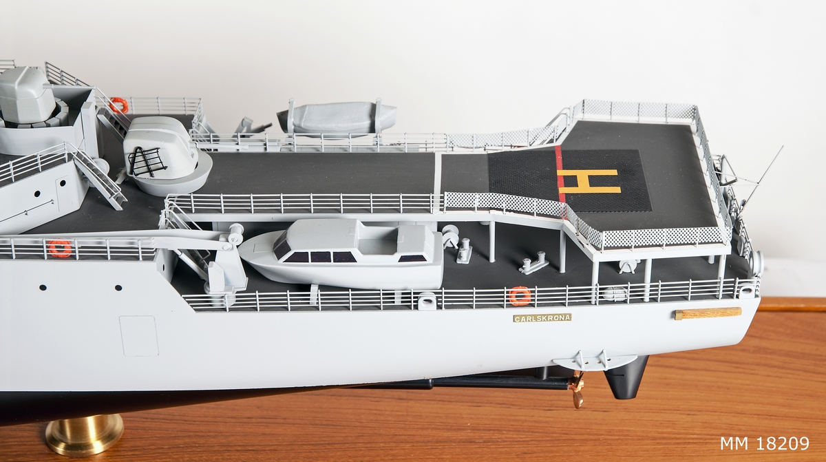 Fartygsmodell av metall och trä föreställande minfartyget Carlskrona placerad i monter. Målad i ljus grått, svart under kölvattenlinjen. Två propellrar i mässing och två roder. Skråvet har två rader ventiler, i fören målat med vitt " M 01 ". Däcket är mörkgrått. Livbåtar, i aktern helikopterplattform märkt med stort orange H. Luftvärnspjäser i för och akter, antenner. Modellen är placerad i monter bestående av rektangulär teakplatta med glashuva och är fästad till plattan med hjäp av två hållare av mässing. På plattan finns två metallplattor med graverad text placerad på vardera kortsida, samma text på båda plattorna. "Minfartyget Carlskrona, leverantör Karlskronavarvet AB, leveransår 1982, Byggt för försvarets materielverk, Längd överallt 105,70 m, Bredd mallad 15,20 m, Djupgåend mallat till KVL 4,00 m, Deplacement till KVL inkl. bihang och bordl. 3138 kubikmeter, Maskinstyrka 4 x 1940 k W."
Monterns mått: H = 420 mm  L = 1170 mm B = 260 mm