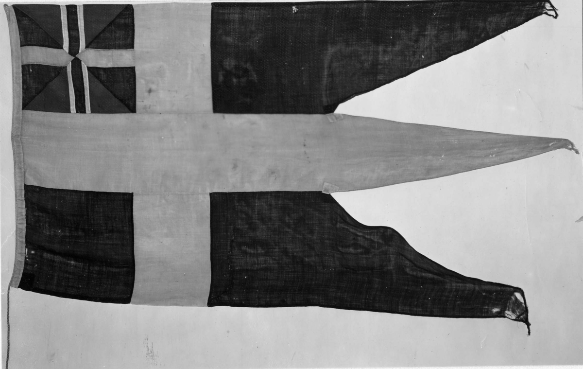 Örlogsflagg, svensk, med yngre unionsmärket. Handsydd av ylleduk med foderduk av linne och lina av sisal. Något sliten och lagad.