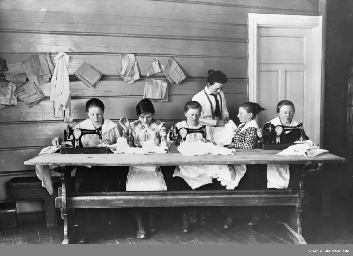Seks personer på sykurs i banken på Lesja i 1919-20. Fem sitter rundt bordet hvorav tre sitter foran en symaskin.