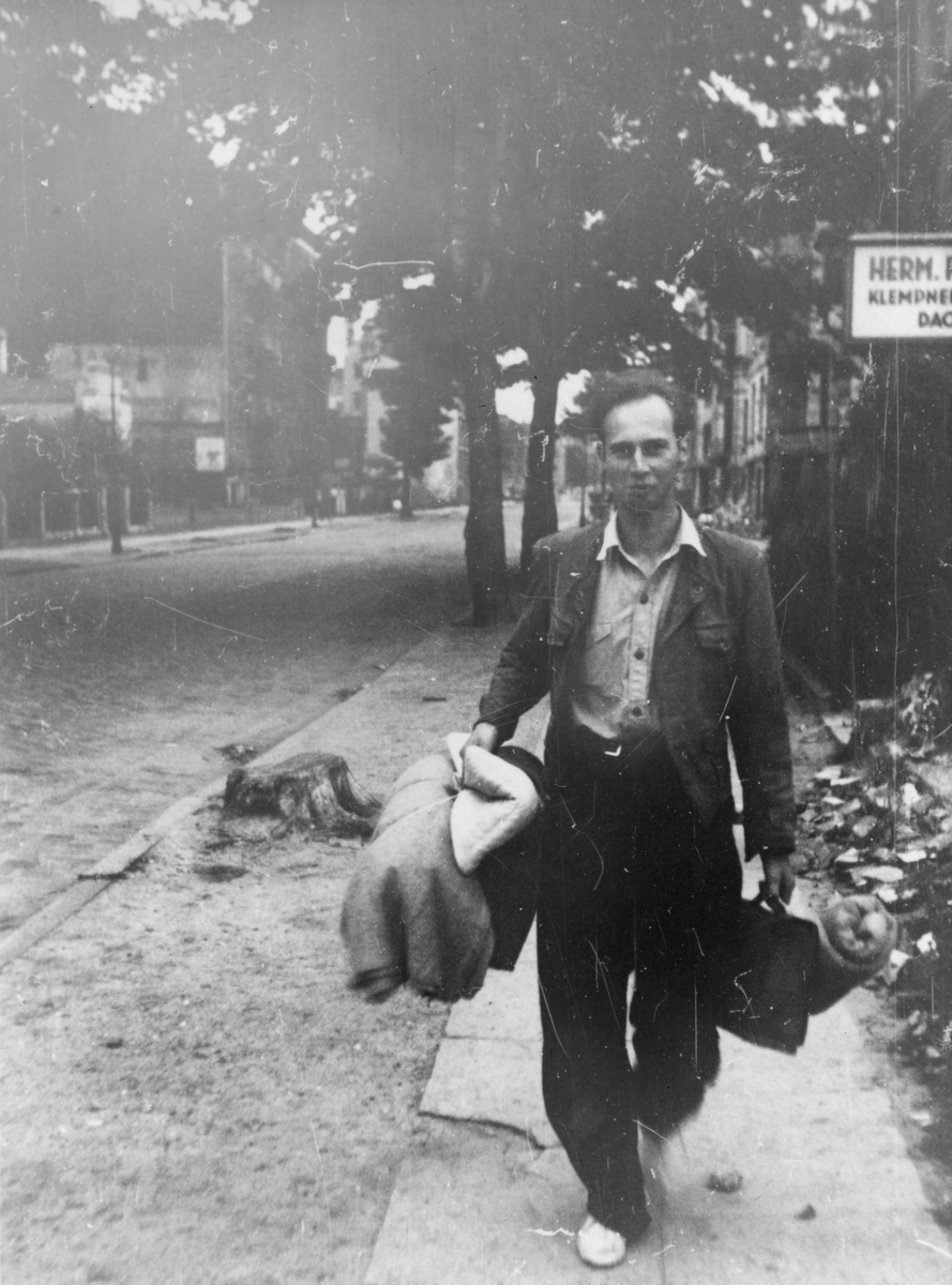Unge Knut Hannestad gående nedover fortau i Hamburg, bærende på sammenrullede tepper og vesker.