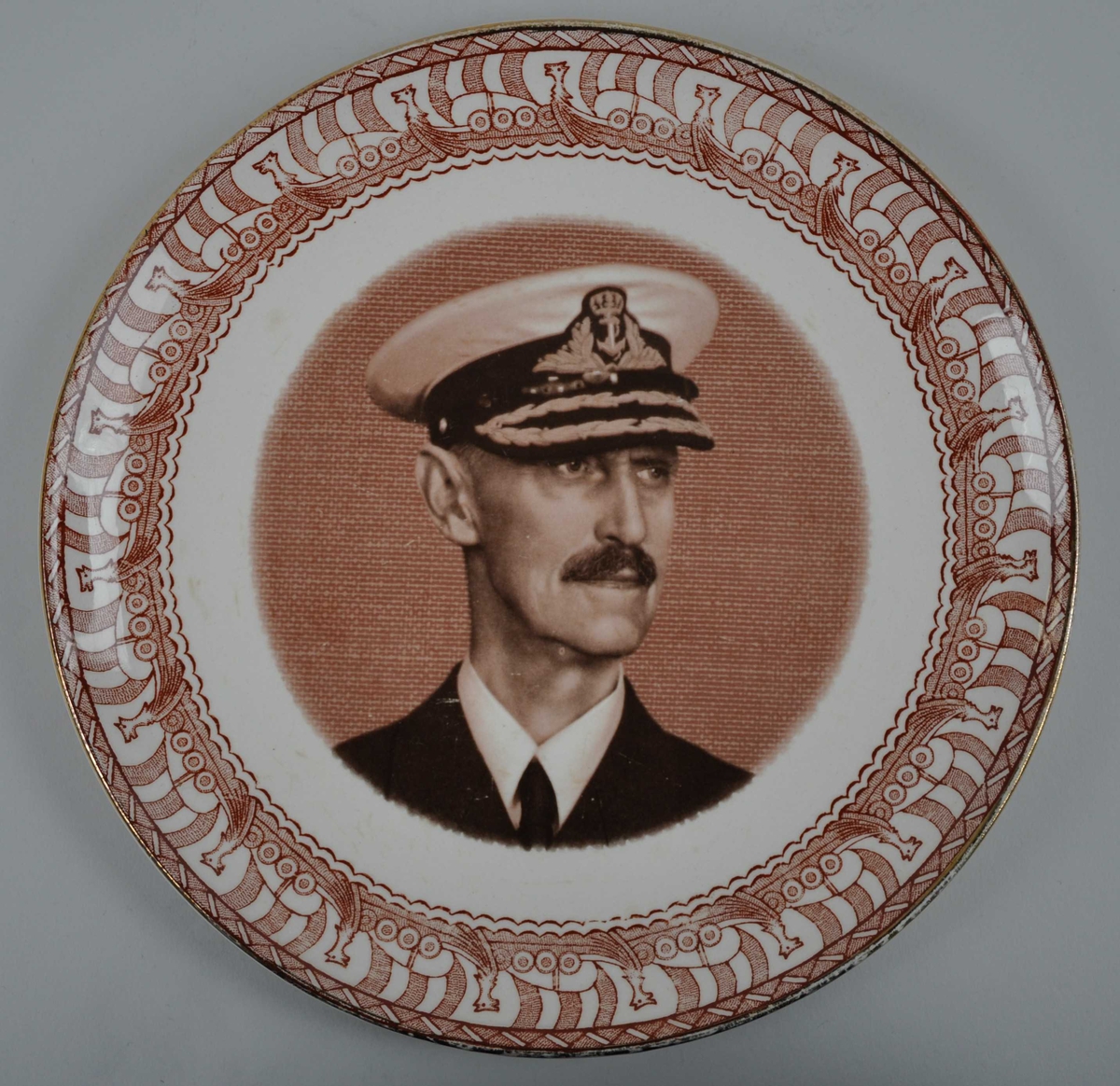 Ein samanhengjande ring med overlappande vikingskip med segl rundt eit portrettet av kong Haakon 7 med marineoffiserslue. 