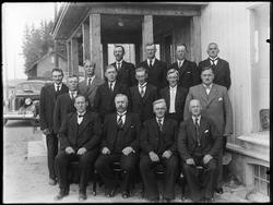 Gruppe menn sittende på en benk utendørs.Noen står, bil i ba