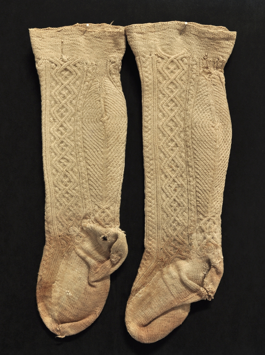 Fra protokollen: Strømper: Materiale: Sauekvitt ullgarn, hjemmespunnet, 3-tråds. Mønster: Øverst 1 cm sjakkbrett ruter av 4 r og 4 vr. Deretter 4 cm rette masker. Skaftene er så mønsterstrikket "vriuband" - med vertikalt betont mønster av fletter og ruter. Legg-partiet er sterkt betont og dekorativt utformet av parallelle rader av vridde rette og vrang-masker, først utover løpende sålenge økningen ved siden av midtmaskene foregår, deretter felling. 
Strømpene er en del hullet. Føttene brun-skjoldet. Hælene masket sammen på undersiden. Hæl og undersiden av foten er påstrikket og det opprinnelige klippet bort.