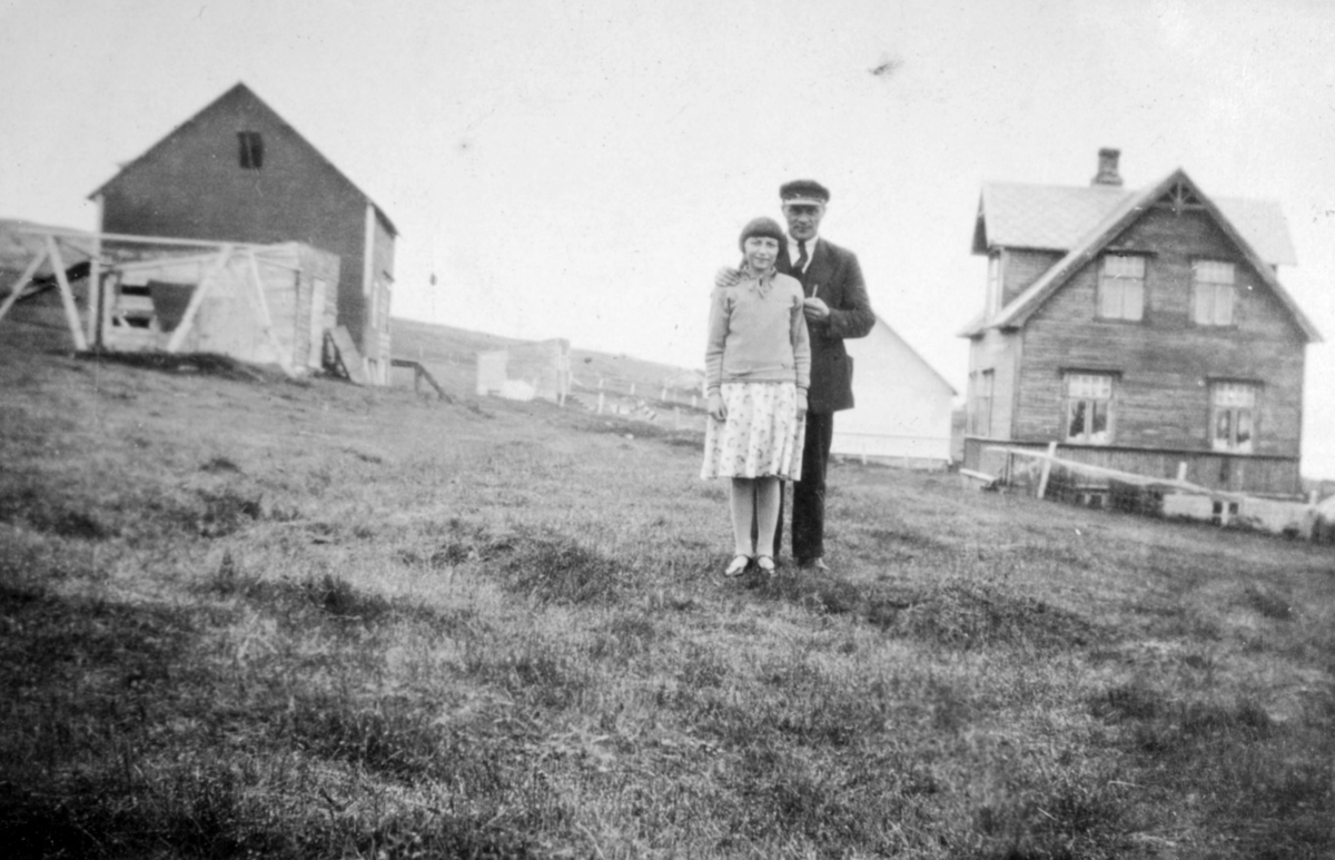 En mann og en jente, muligens far og datter, poserer for fotografen på et jorde. Flere bygninger ses i bakgrunnen. Sted og personer er ukjent, men bildet kan være tatt i Kvalsund kommune.