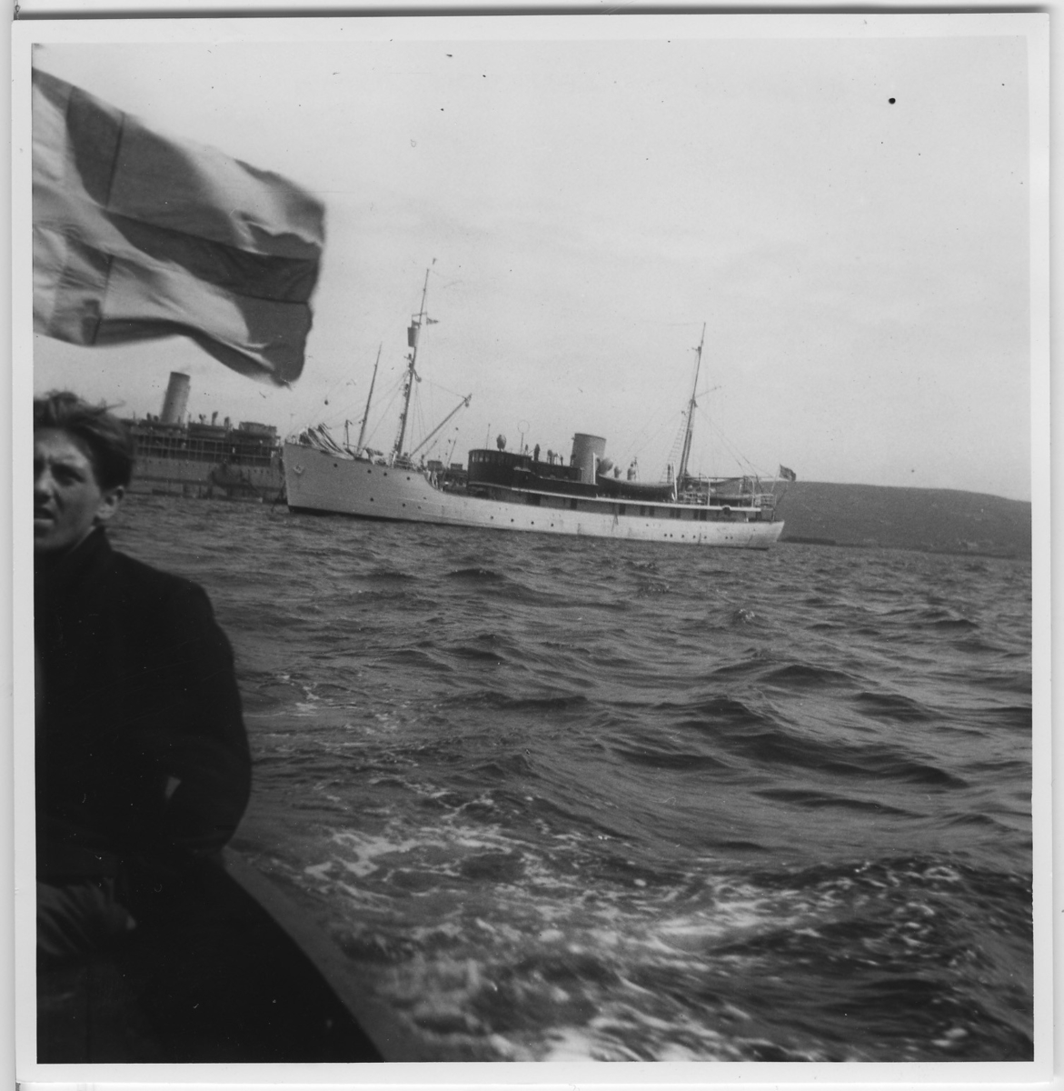 'Från ''Skageraks''-expeditionen till Hebriderna och Shetland: ::  :: ''Skagerak i Lerwicks hamn, Man vid flagga på annan båt i förgrunden (förstorad version av foto nr 4113:27) ::  :: Ingår i serie med fotonr. 4113:1-82.'