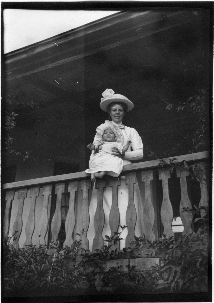 'Diverse fotografier från bl.a. dåvarande Nordrhodesia, nu Zambia, tagna av Konsul Magnus Leijer. ::  :: En kvinna ståendes på en veranda vid räcket, på räcket håller hon en sittande, skrattande bebis.'