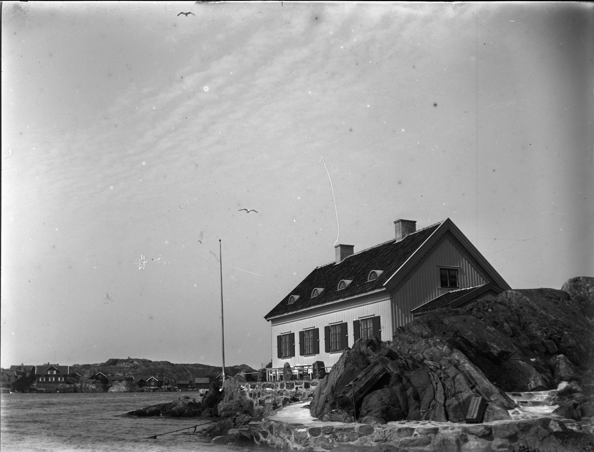 'Bildtext: ''Algott och Hjordis.'' :: Vy med ett hus liggande på klippor ända ner vid vattnet, murad stenmur. Vy med havet och på avstånd hus. ::  :: Ingår i serie med fotonr. 5233:1-14'