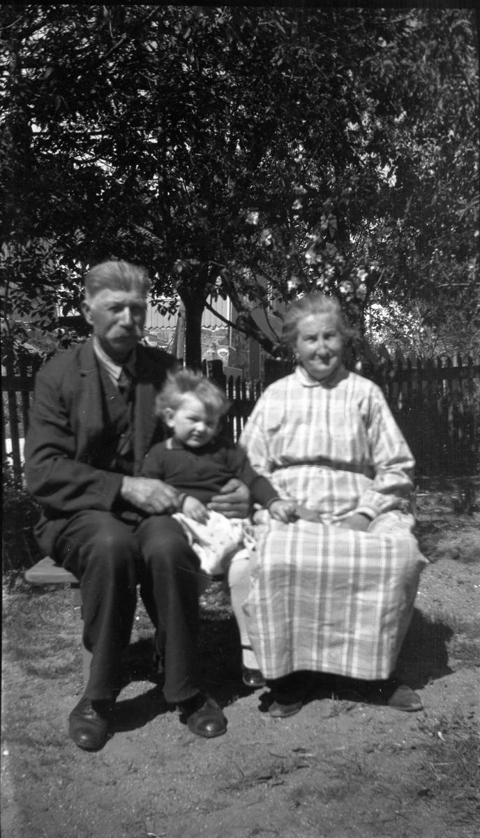 'Gruppbild med 1 man, 1 kvinna och ett litet barn sittande på träbänk i trädgård. I bakgrunden ett trästaket. Klädsel. ::  :: Ingår i serie med fotonr. 5233:1-14'