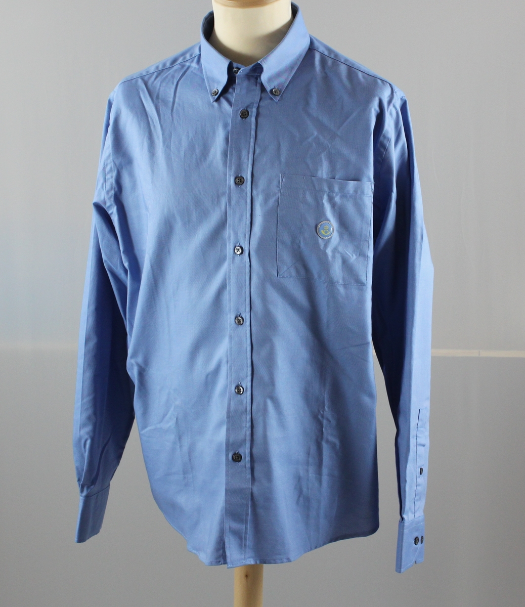 Skjorta i ljusblått med långa ärmar. En bröstficka till vänster och ett reflexmärke med Postens symbol på den. Märkt "herr" och storlek 40.