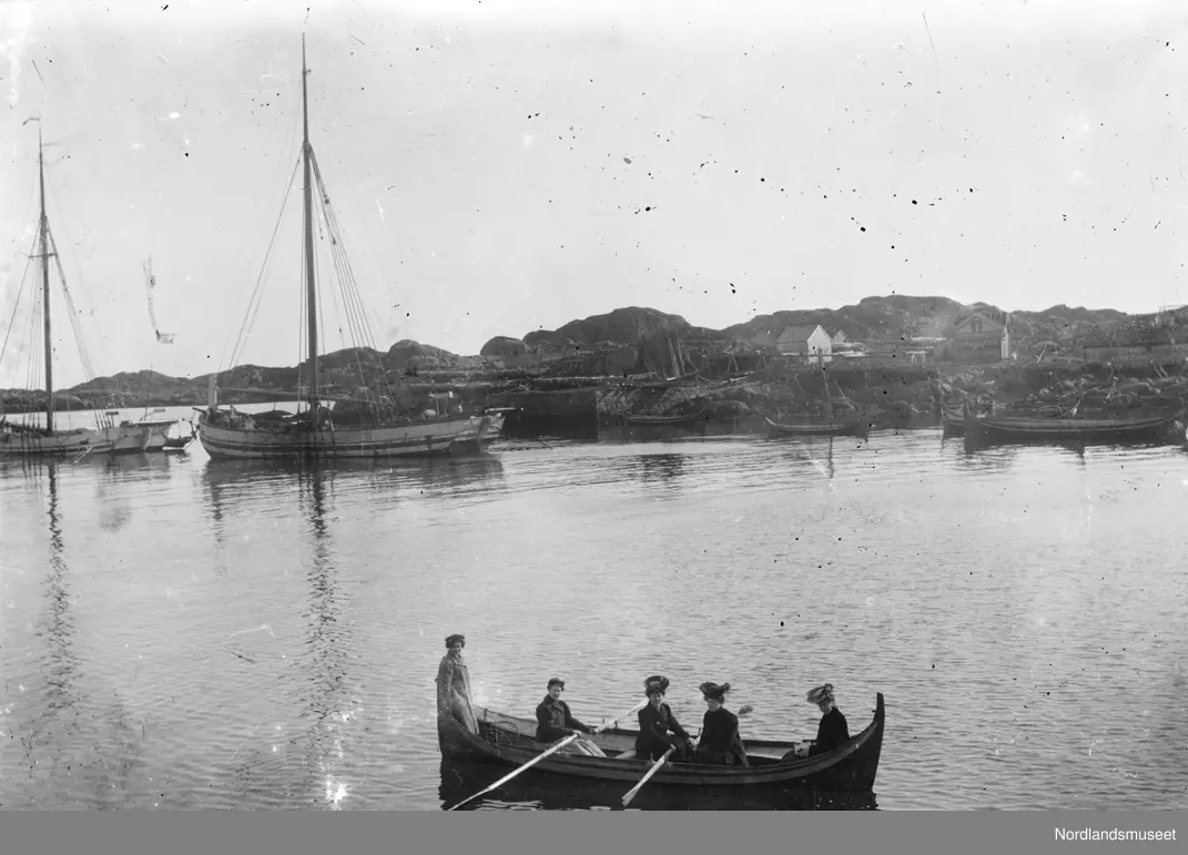 Fem kvinner som ror i en båt, flere store båter ligger på havnen. Inne ...