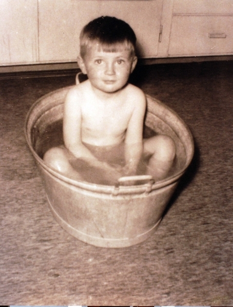 Badestamp i kjøkkenmiljø. Gutt, kjøkkenskap, skuffer. 1-2 år gammel gutt sjitter i badestamp, sett fra  stampens kortside. Linoleumsgulv, i bakgrunnen  nedre del av lysmalte kjølkenskap og skuffer.