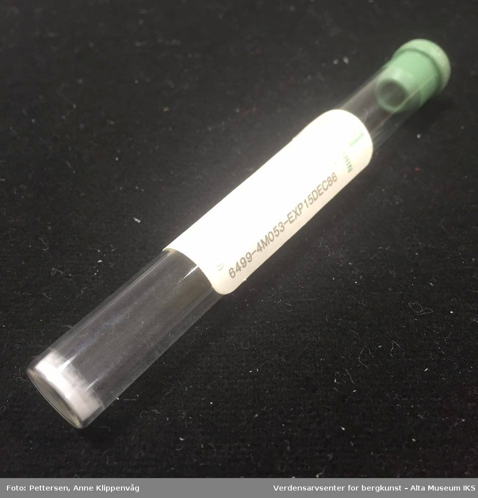 En sylindrisk beholder av glass, med grønn gummikork. I bunnen ser man et lite lag av hvitt pulver.