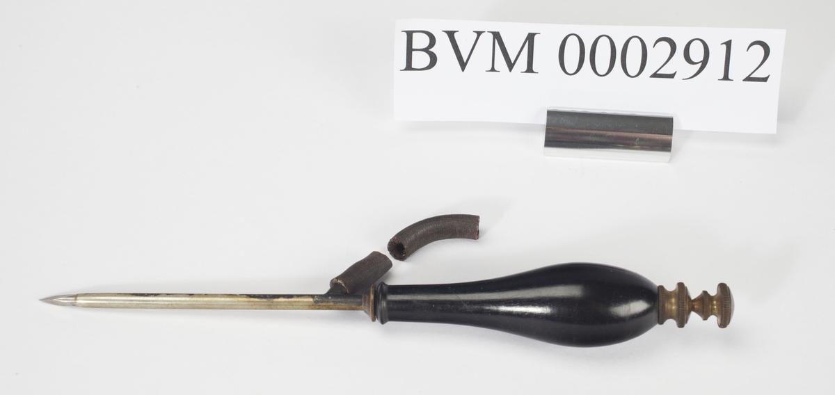 Syllignende instrument med av stål med svart lakkert treskaft. Øverst mot håndtaket et siderør av gummi. En liten del av gummirøret er knekt av.