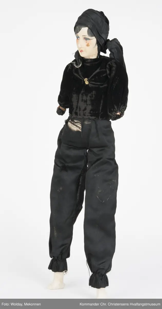 Dukken: sminket kvinneskikkelse med sigarett i munnen,  tørkle på hodet og sort todelt dress (Langermet, kort bluse, lange bukser, strømper.