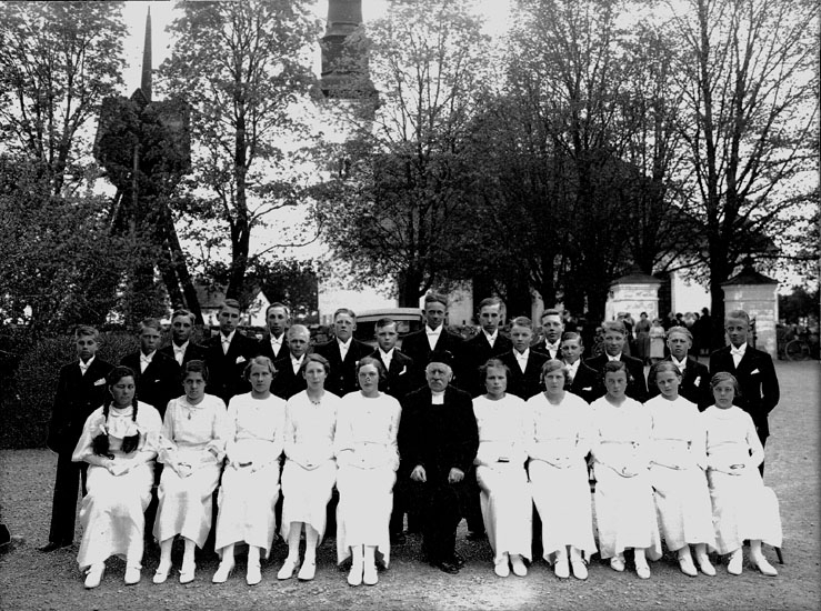 Konfirmander, 10 flickor, 16 pojkar och kyrkoherde Ivar Jäderlund.
Glanshammars kyrka och klockstapeln i bakgrunden.