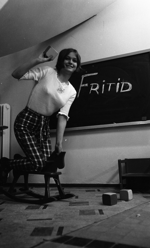 Modebilder 23 april 1966

En ung kvinna står på knä på en gunghäst iklädd vit blus, rutiga byxor och svarta lågklackade skor. Hon skrattar och håller en lekkolss i sin högra hand. Det är en modebild. Bakom henne finns en rittavla där någon skrivit ordet "Fritid" med krita. Lekklossar och en liten låg barnstol står på golvet bl.a.