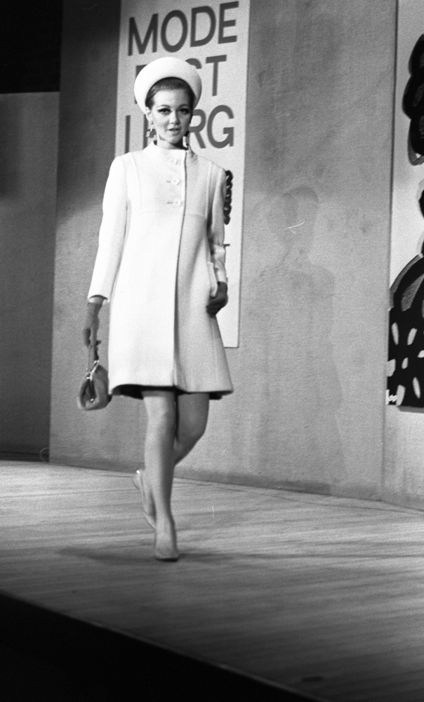 Modevisning, Görtz motor (Rep av oil) 6 april 1967

En fotomodell går på scenen under en modevisning. Hon är klädd i en vit kappa, vit hatt, örhängen, pumps på fötterna, handskar och hon håller en handväska i höger hand. Bakom henne på väggen hänger en skylt med texten: "Modefest i färg varuhuset Domus."