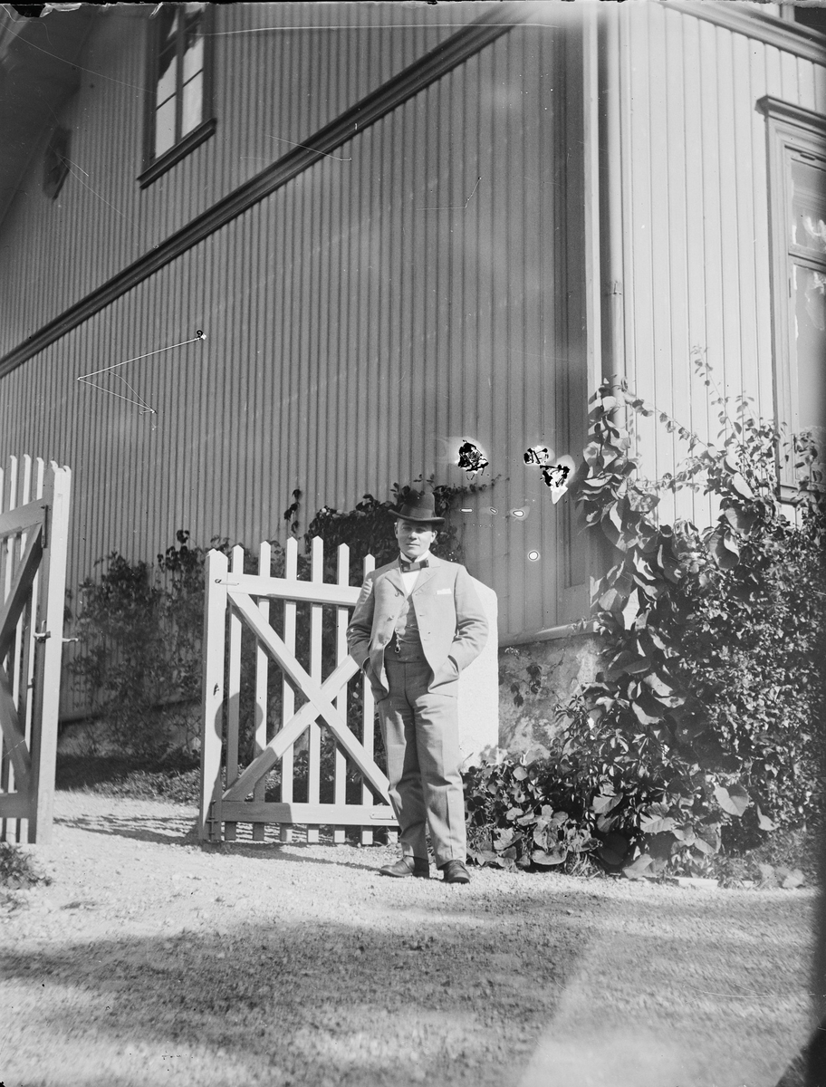 Christian Pierre Mathiesen står ved en åpen port. Bak ham ligger et hus med klatreplanter langs veggene. Solen skinner.