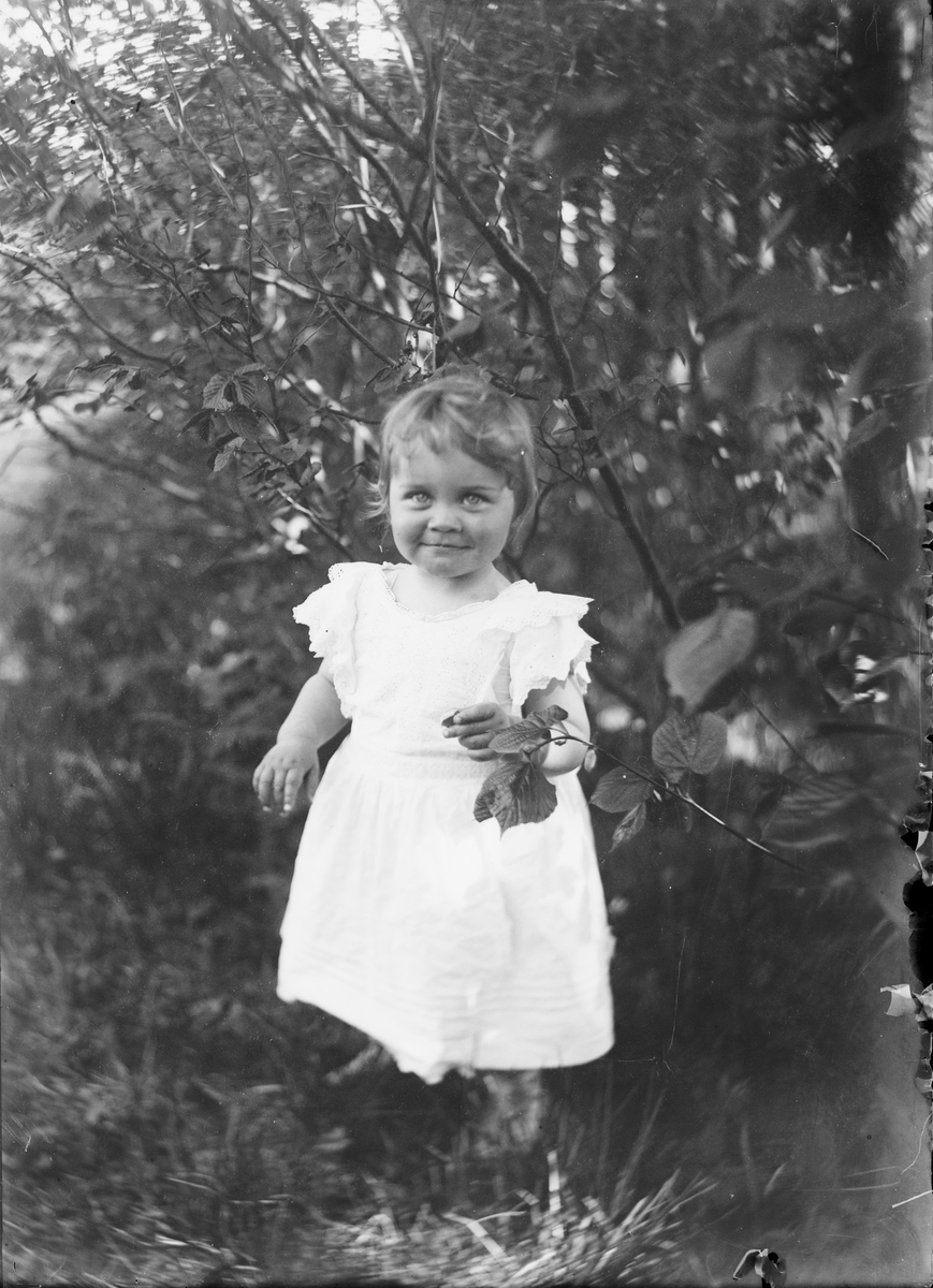 Et barn i står i gresset, ved busker og trær, og smiler til fotografen.