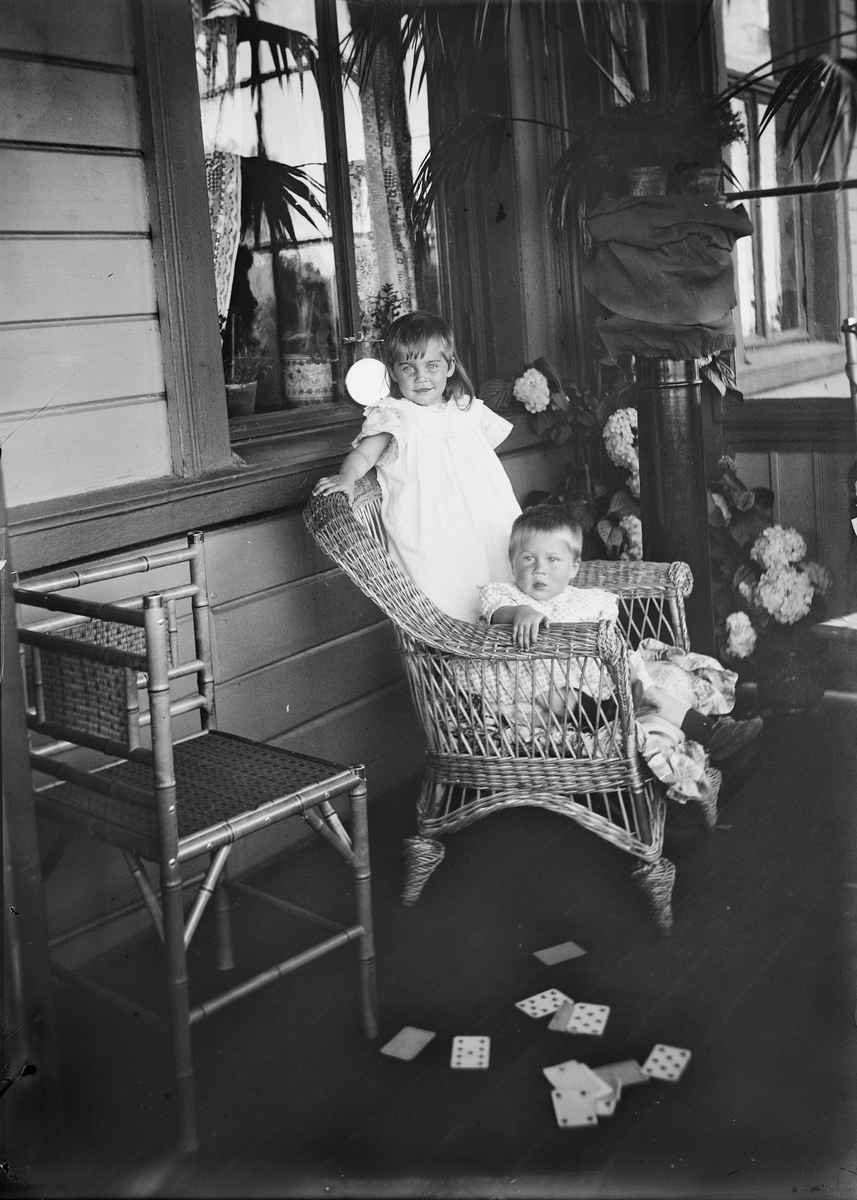 En gutt sitter i en kurvstol og bak ham i stolen står en liten jente. De er fortografert på en veranda. På bakken ligger en kortstokk. I bakgrunnen sees vinduer inn til huset og blomstrende hortensiaer. I forgrunnen sees en stol.