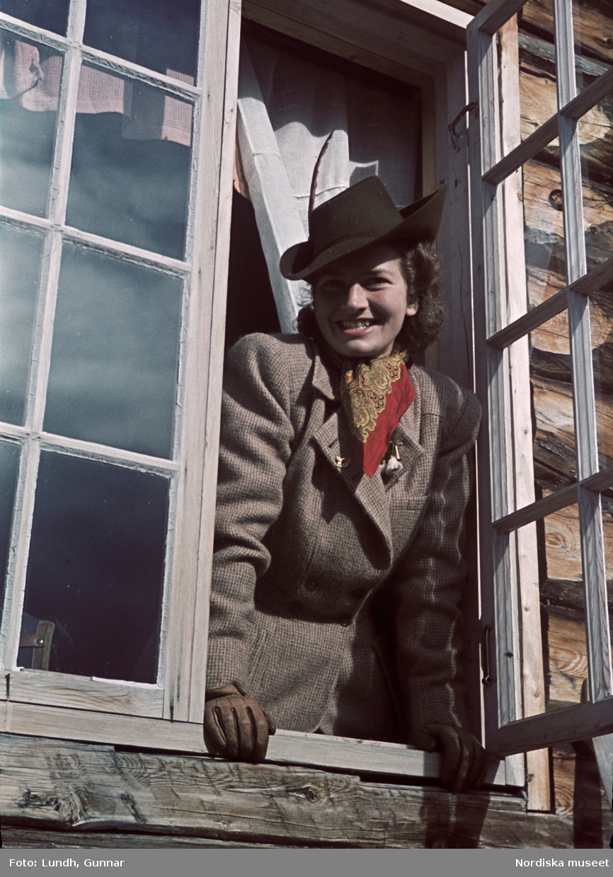 Tweedeklädd kvinna i hatt blickar ut genom ett öppet fönster.