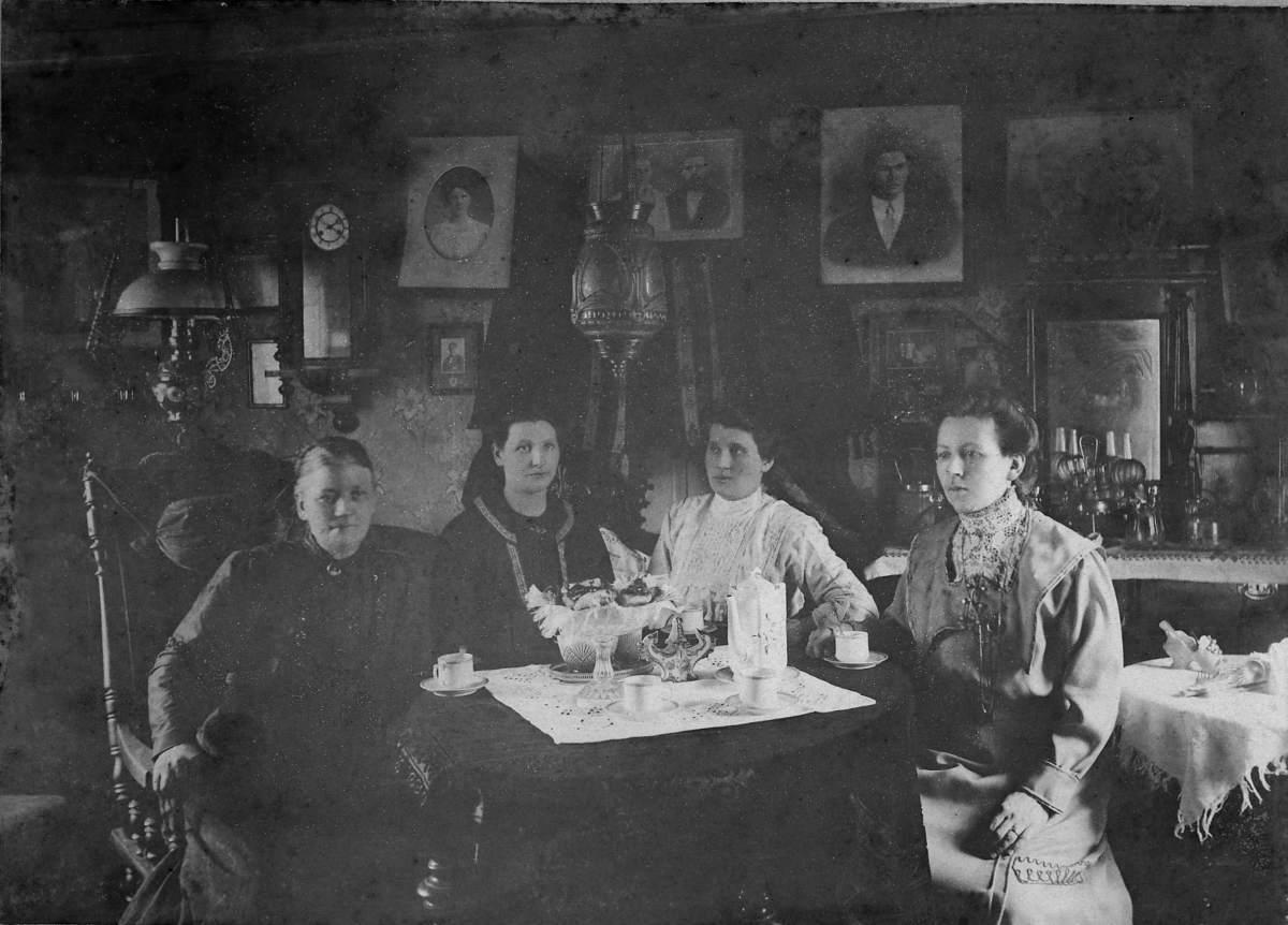 Fire damer sitter i stue til Kima og Peder Lein i Berlevåg.  Fra venstre: Kima Lein, hennes dødre Astrid g. Larsen og Emma g. Mathisen, og svigerdatter Emma Lein f.Wicander. Kvinnene sitter ved et kaffebord. På bordet porselenkopper, kaffekanne og fat med kaker. Emma Lein f. Wicander var født i Syltefjord, dro til Amerika 1901 og giftet seg der med Kimas søn Enok Lein. Familien kom tilbake og bosatte seg i Berlevåg/Båtsfjord i 1911.  Kjolen hennes er tydelig fra denne emigrasjonen.