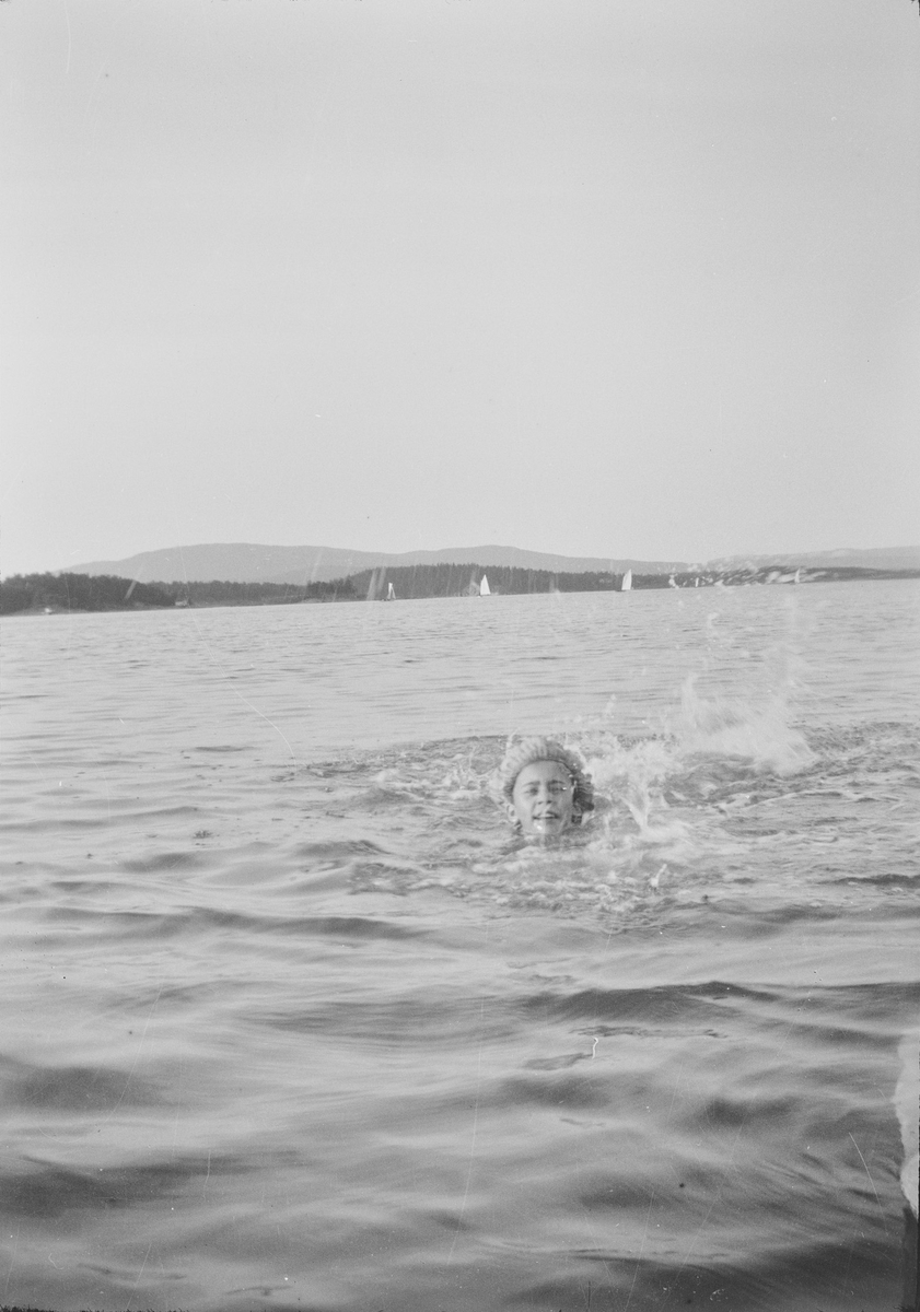 Celina Marie svømmer i en fjord med badehette på. I bakgrunnen sees seil fra små seilbåter/fritidsbåter.