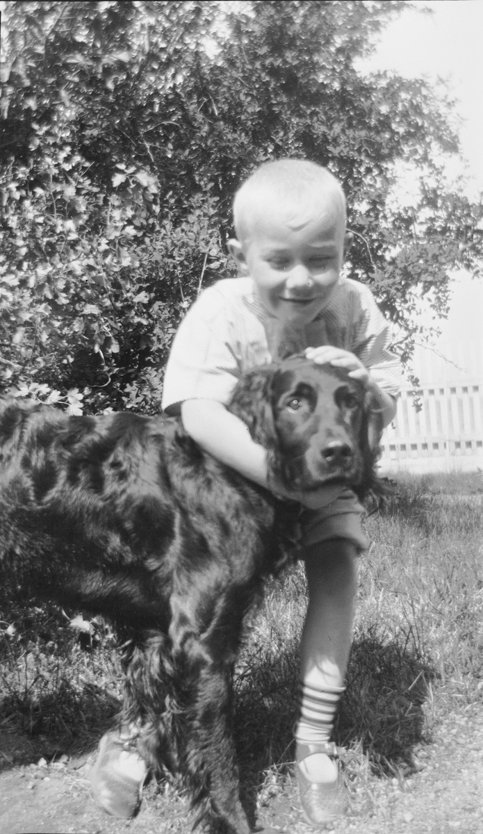 En smilende Iacob Ihlen Mathiesen står i en hage og klapper en hund på hodet, trolig en Gordon setter. Bak hunden og gutten sees hagetrær og et stakittgjerde. Det er sommer.