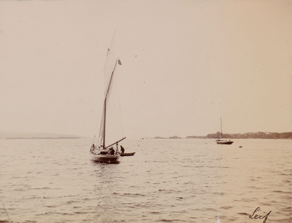 Seilbåten "Leif" med flere mennesker ombord, muligens er mannen bakerst i båten Haaken Larpent Mathiesen. I robåten ved siden av står en mann og fisker kanskje.