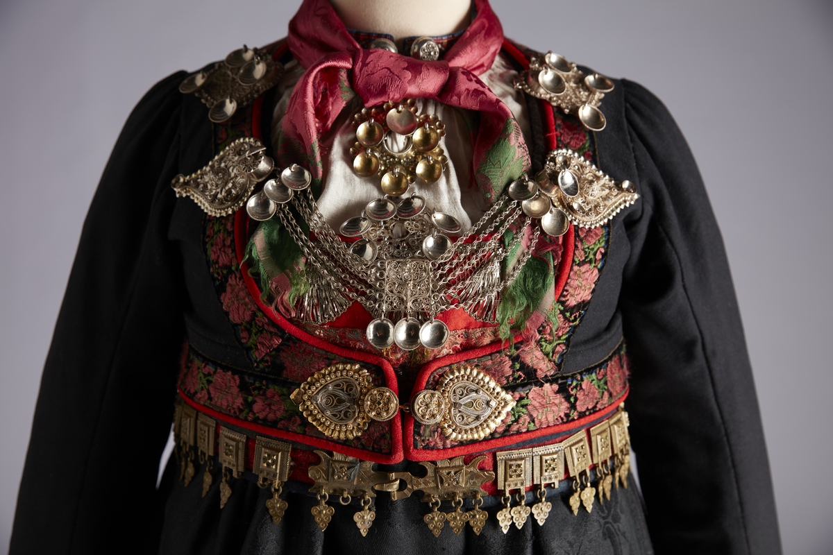Forkle i svart ulldamask med kanting av rødt klede med sølvknipling og brosjert silkebånd med blomstermønster.