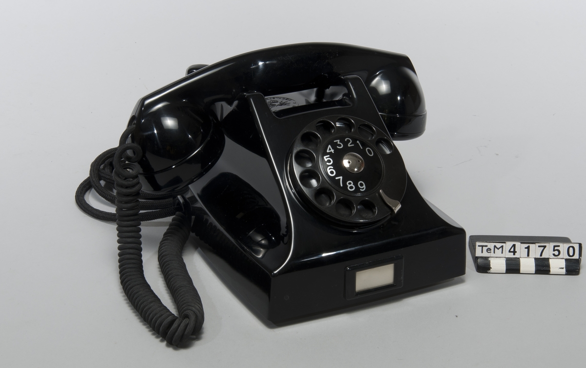 Telefonapparat BC 560, ändstationsapparat för AT-system. Bordapparat modell m50 av svart bakelit med apparatsnöre anslutet till väggplint med lock av vit plast.