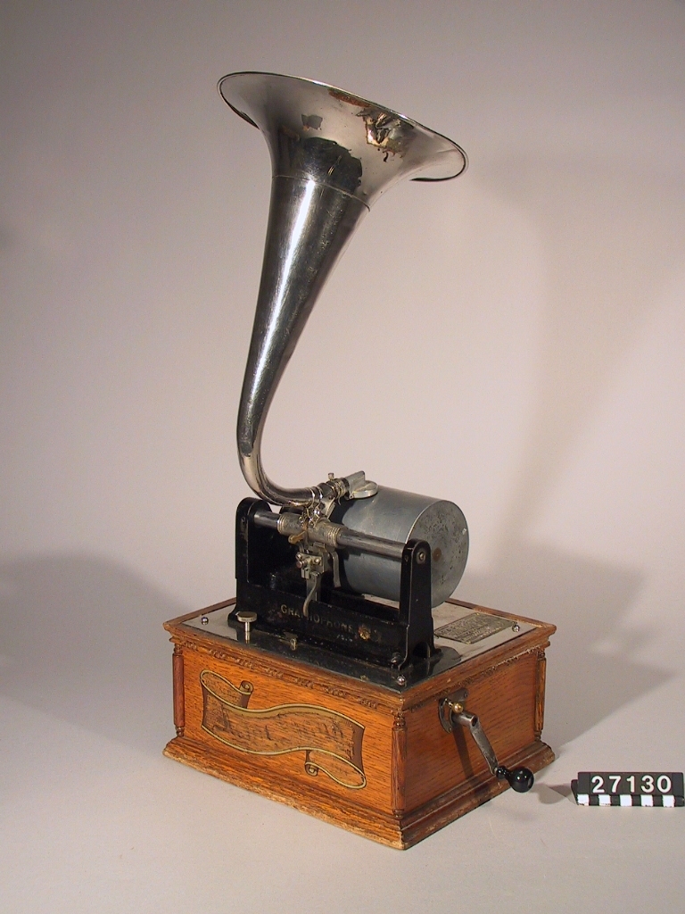 Fonograf, med svarvanordning. Avsedd för 5 tums vaxrullar, 120 varv/minut. Skylt: "The Graphophone patented May 4 1886 .... Mar 30 1897 Manufactured by the American Graphophone Company, New York, N.Y. Type HG 1216".
Tillbehör: Lösa delar: apparat, vev, ljuddosa för återgivning, ljuddosa för upptagning (båda i pappask), böjd tratt, huv av trä samt ytterhuv av brunt läder.