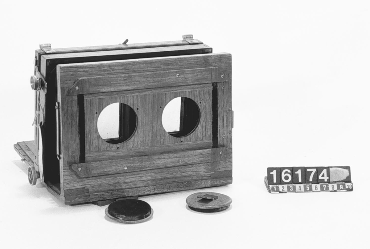 Sterokamera av trä, för tagning av bilder 8.5 x 8.5 cm, med ett skjutbart objektiv. Objektiv med bräde saknas.
Tillbehör: 2 st. lock till hålen för objektiven.