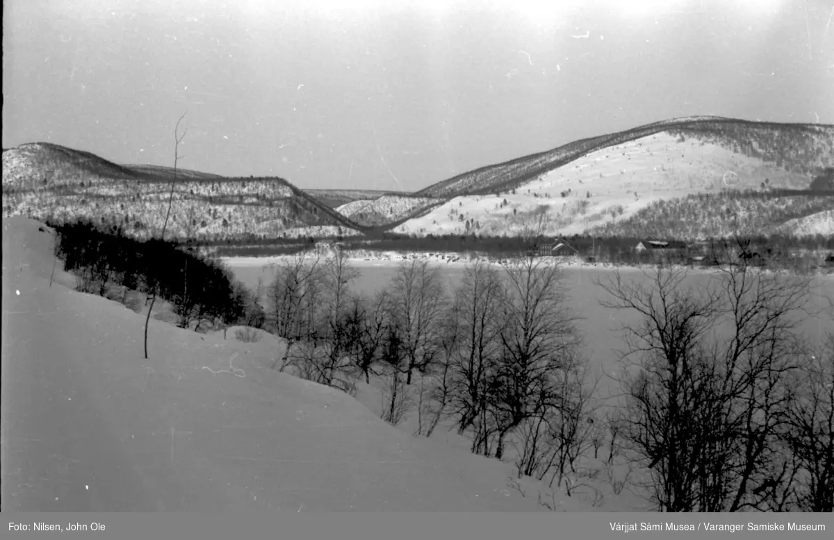 Snødekt vei, frosen elv eller vann, hus i bakgrunnen. Ukjent sted 23. april-1966