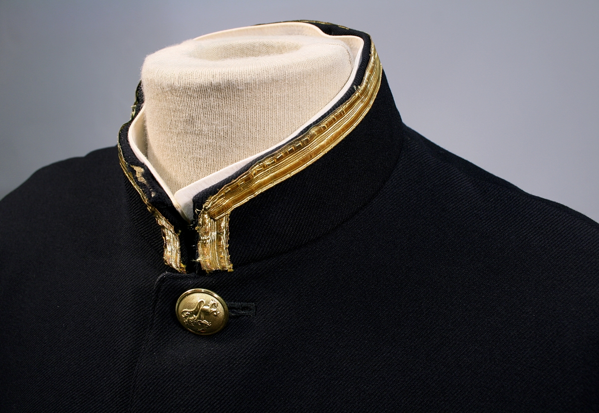 Uniformsjakke i sort med blåskjær. Enkeltspent med seks forgylte riksløveknapper. Stående krage kantet med 12 mm gullbånd. To brystlommer.