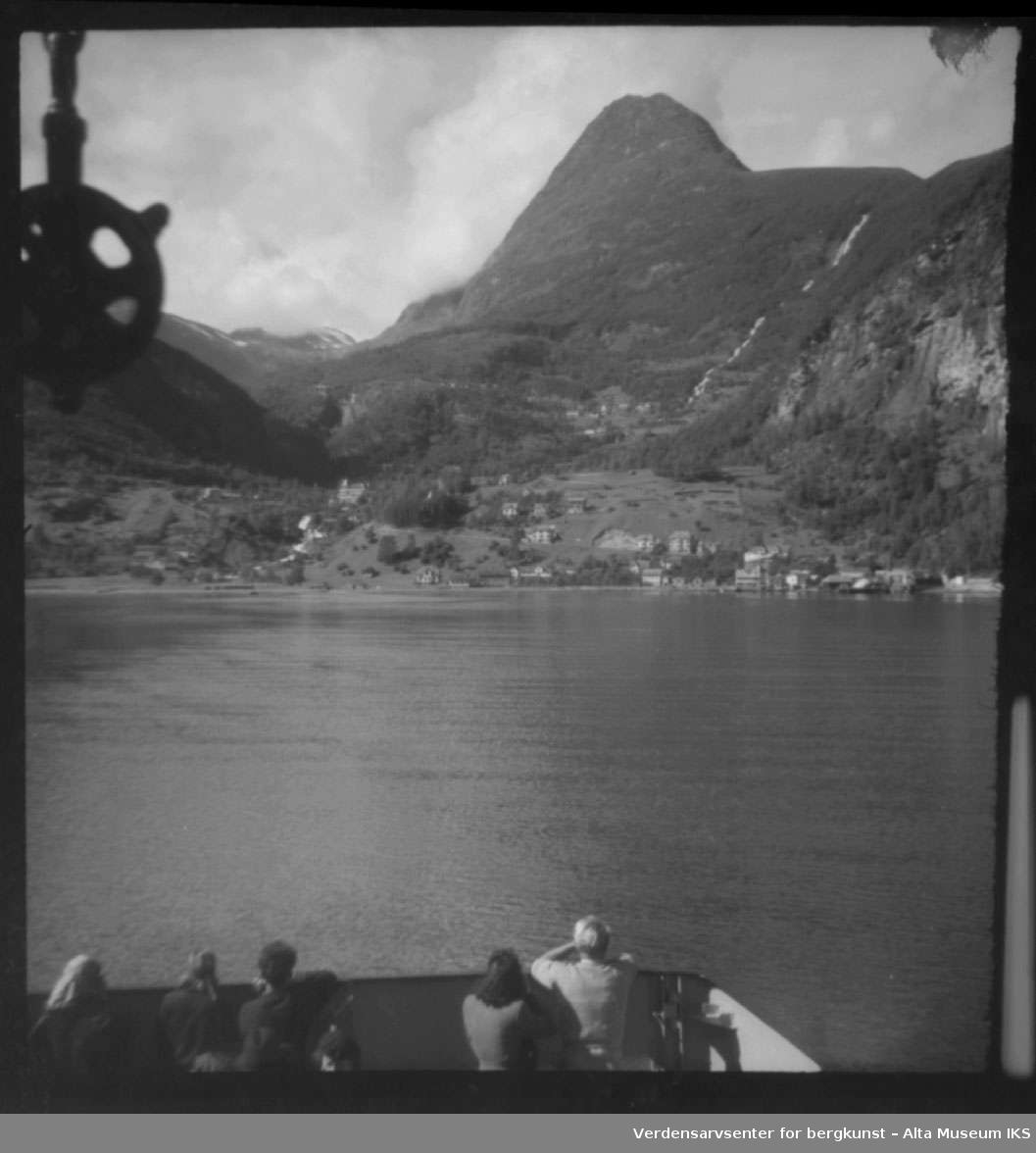Utsikt over fjord fra båt, på land ser vi en bygd med flere hus/bygninger og høye fjell.